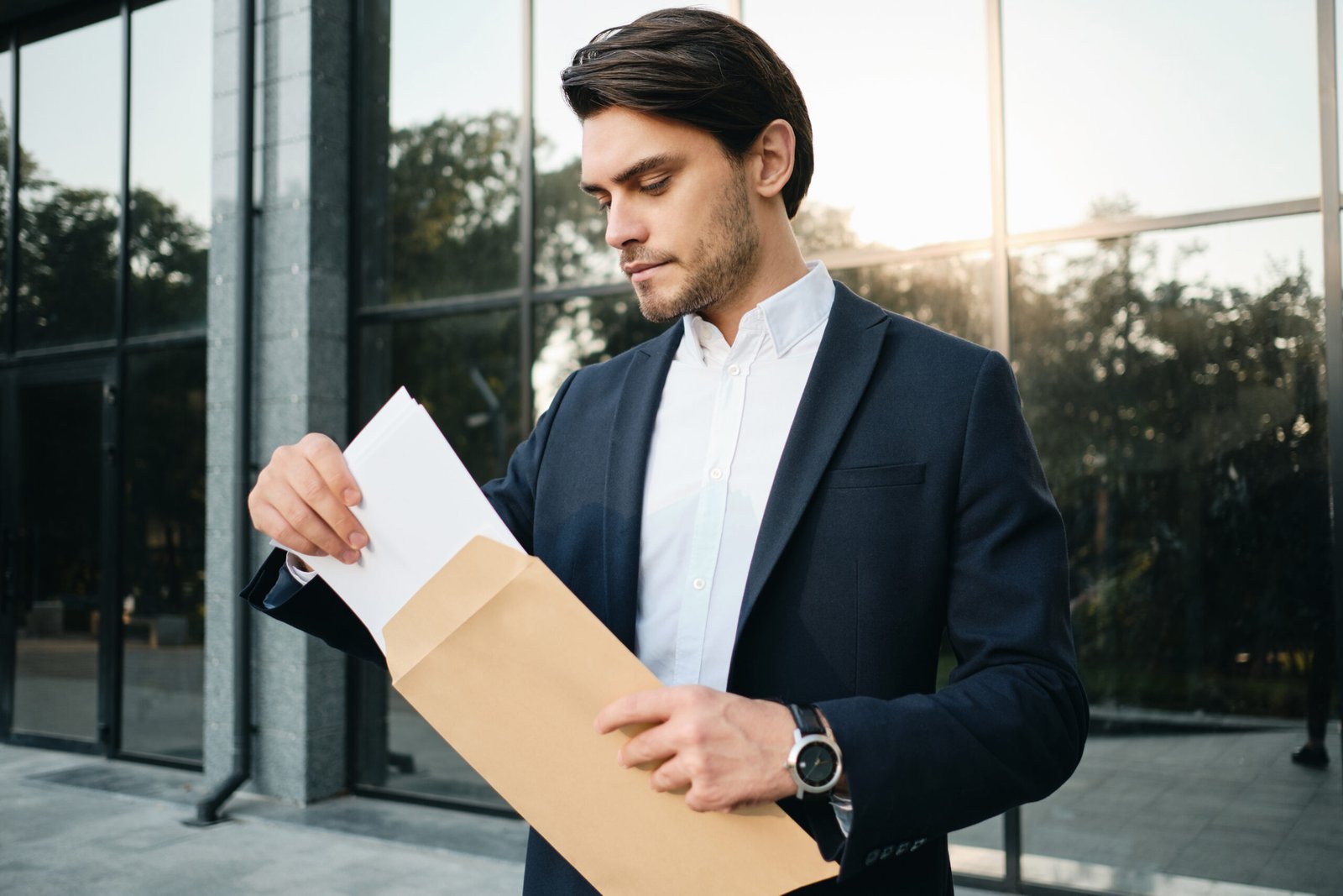 Young handsome bearded brunette man in white shirt and classic jacket standing and dreamily opening envelope with glass building on background